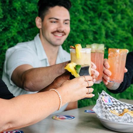 Group of friends clinking colorful tropical cocktails at an outdoor bar with a green hedge wall, pineapple garnishes and a basket of chips — summer happy hour vibe