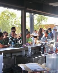 Sunlit outdoor bar patio with a lively crowd gathered along the bar counter, people chatting over beers and cocktails, beer taps, string lights and trees visible outside.