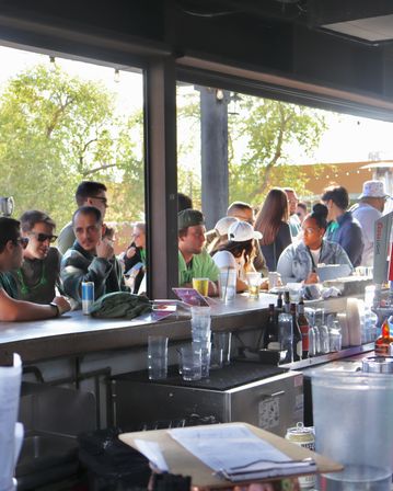 Sunlit outdoor bar patio with a lively crowd gathered along the bar counter, people chatting over beers and cocktails, beer taps, string lights and trees visible outside.