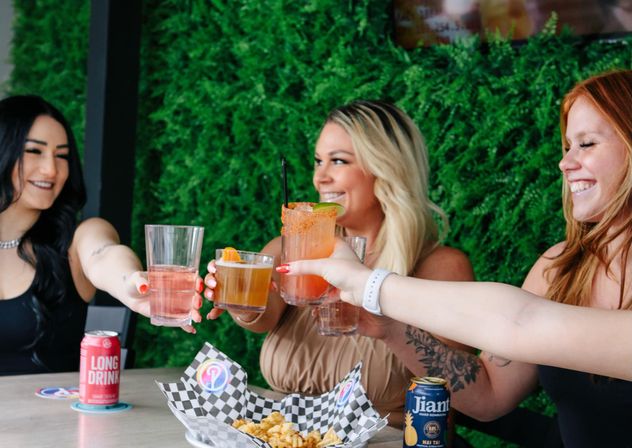 Three smiling women clinking cocktails and beer cans over a table with a basket of fries, seated at an outdoor patio with a lush green plant wall.
