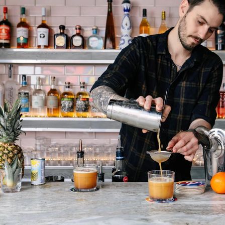 Tattooed bartender pouring shaken craft cocktails through a strainer into short glasses on a marble bar counter, with backlit liquor shelves, pineapple and orange nearby.