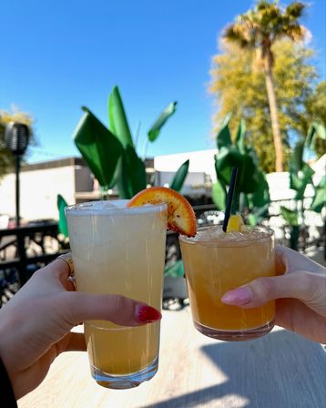 Two citrus cocktails raised for a toast on a sunny outdoor patio — iced pale-yellow drinks with orange garnish, tropical plants and a palm tree in the background, bright brunch vibes.