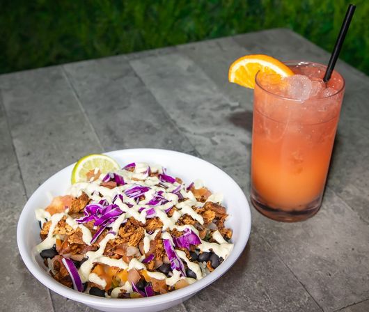 Colorful Tex-Mex bowl with seasoned shredded protein, black beans, pico, shredded purple cabbage, creamy white drizzle and lime wedge, paired with an iced pink-orange cocktail garnished with an orange slice on a concrete patio table.