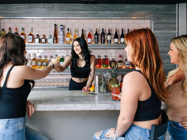 Smiling bartender hands a cocktail to a customer at a modern marble-topped bar with backlit liquor shelves, a fresh pineapple on the counter, and three friends laughing over drinks.