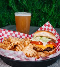 American cheeseburger with melted cheddar, tomato and lettuce served with crispy waffle fries in a red-check paper basket and a pint of beer on an outdoor patio table