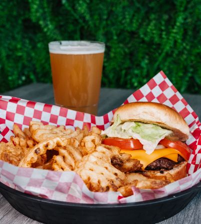 American cheeseburger with melted cheddar, tomato and lettuce served with crispy waffle fries in a red-check paper basket and a pint of beer on an outdoor patio table