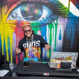 DJ in sunglasses and cap at an urban pop-up booth operating a controller and sticker-covered laptop in front of a vibrant dripping rainbow-eye street art mural.