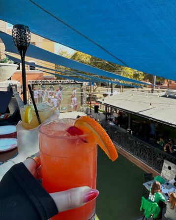 Bright orange cocktail with orange slice and cherry and a lemon-garnished drink raised on a sunny rooftop sports-bar patio under blue shade sails, overlooking a large outdoor screen showing a football game and fans below.