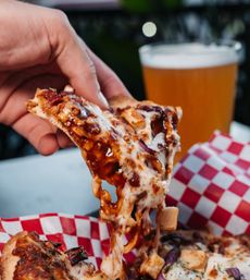 Hand lifting a gooey BBQ chicken pizza slice with melted cheese, red onion and barbecue sauce over red-and-white checkered paper, pint of beer in the blurred outdoor background.