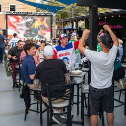 Outdoor sports bar patio with tailgate-style energy — fans cheering at a large outdoor screen showing a football game, high tables with drinks and food, one fan wearing a Montréal jersey.