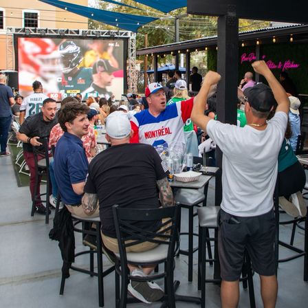 Outdoor sports bar patio with tailgate-style energy — fans cheering at a large outdoor screen showing a football game, high tables with drinks and food, one fan wearing a Montréal jersey.
