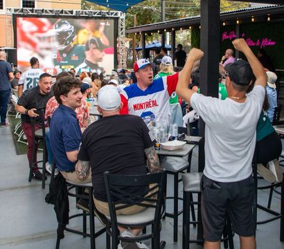 Outdoor sports bar patio with tailgate-style energy — fans cheering at a large outdoor screen showing a football game, high tables with drinks and food, one fan wearing a Montréal jersey.