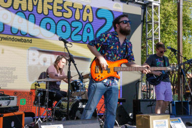 Three-piece band playing an outdoor summer festival stage — bearded bassist in a colorful shirt with an orange electric bass front-center, drummer behind a black kit and guitarist at right against a colorful LED backdrop and trees.