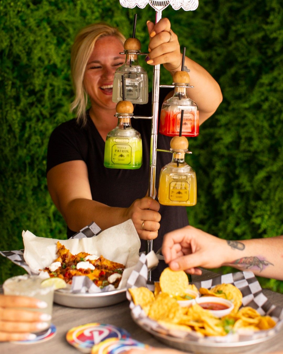 Outdoor patio scene: server presenting a vertical tequila bottle margarita tower as friends reach for chips, salsa and loaded nachos.