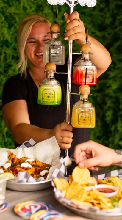 Outdoor patio scene: server presenting a vertical tequila bottle margarita tower as friends reach for chips, salsa and loaded nachos.
