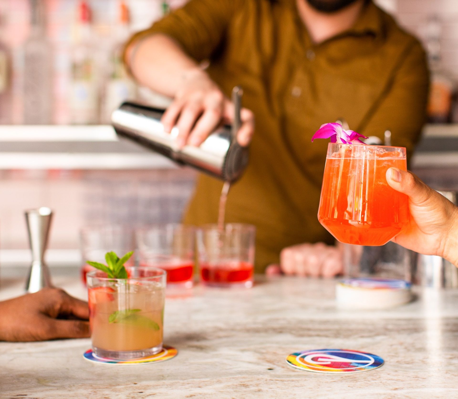 Bartender pouring from a shaker as a hand holds a bright coral cocktail garnished with a purple orchid on a bar counter