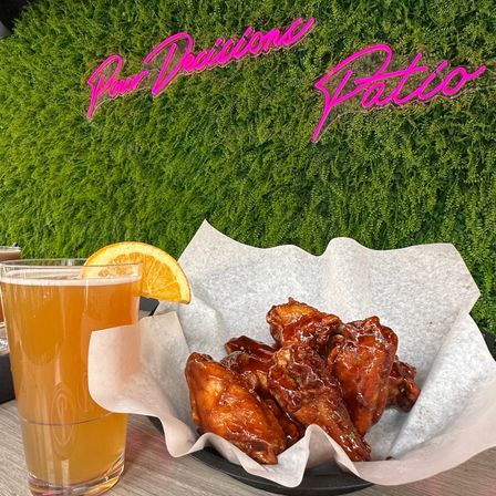 Saucy glazed chicken wings in a paper-lined basket with a pale beer garnished by an orange slice on a patio table, neon pink sign and green plant wall backdrop.