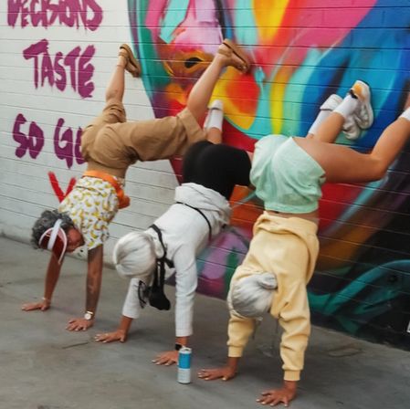 Three women doing handstands against a vibrant graffiti brick mural featuring a colorful abstract face, wearing casual summer clothes and sneakers