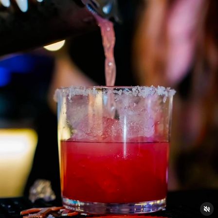 Close-up of a vibrant red cocktail being poured into a salt-rimmed rocks glass over crushed ice, with colorful bar lights blurred in the background