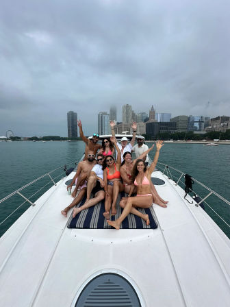 Group of friends lounging and waving on the bow of a white yacht with the Chicago skyline and Navy Pier Ferris wheel across Lake Michigan under a cloudy sky
