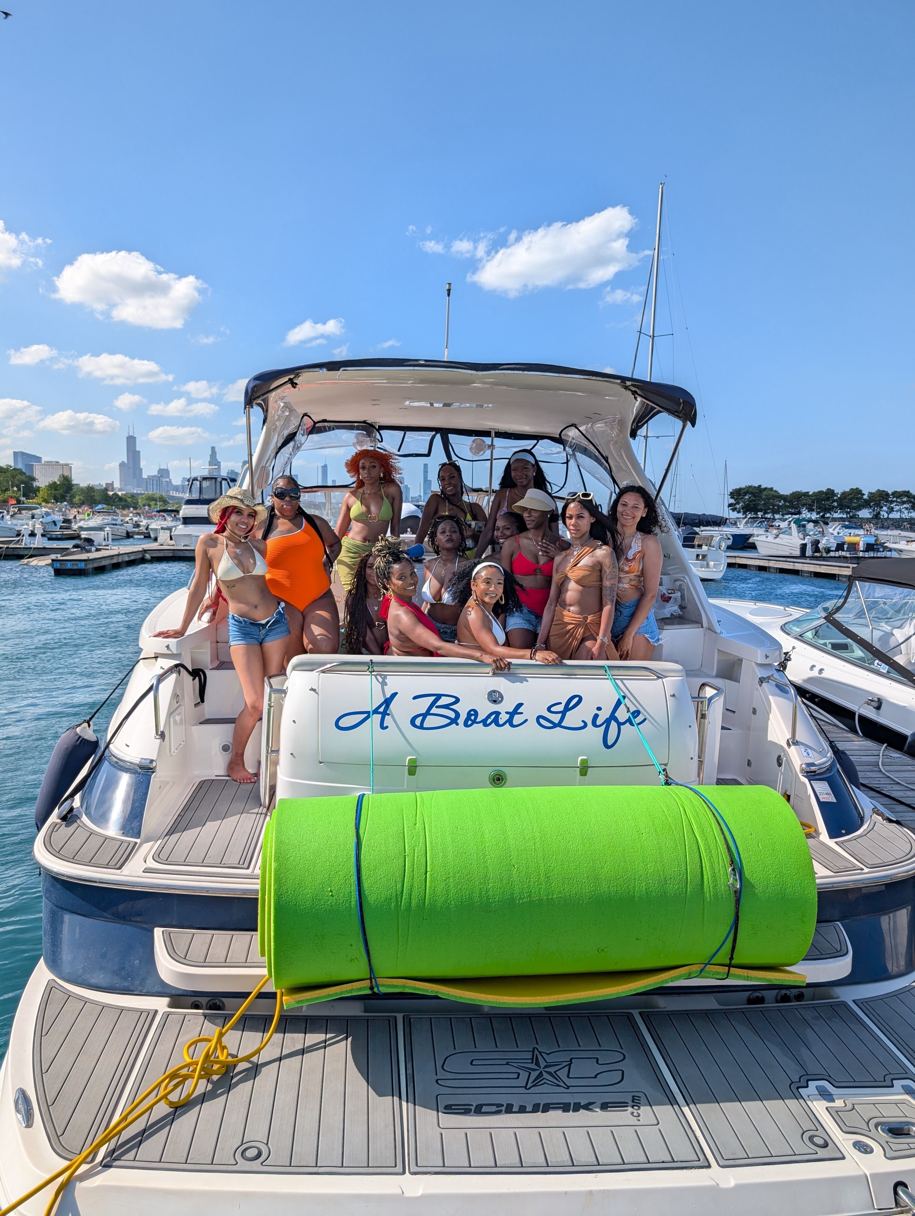 Group of women in colorful swimsuits posing on the stern of a white yacht with a bright green foam roll, docked at a busy marina with a city skyline and clear blue summer sky.