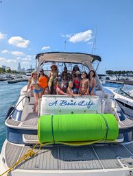 Group of women in colorful swimsuits posing on the stern of a white yacht with a bright green foam roll, docked at a busy marina with a city skyline and clear blue summer sky.