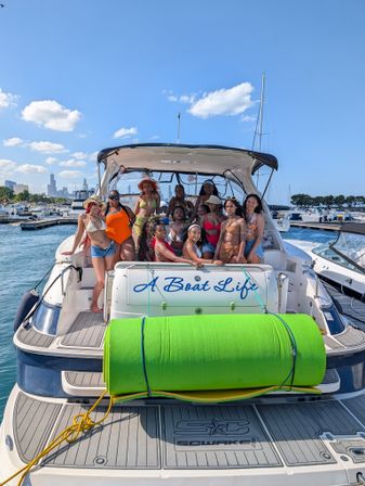Group of women in colorful swimsuits posing on the stern of a white yacht with a bright green foam roll, docked at a busy marina with a city skyline and clear blue summer sky.