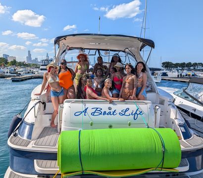 Group of women in colorful swimsuits posing on the stern of a white yacht with a bright green foam roll, docked at a busy marina with a city skyline and clear blue summer sky.