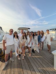 Group of friends in white and neutral summer outfits posing on a wooden marina dock in front of a docked boat, waterfront skyline and wispy blue sky at golden hour.