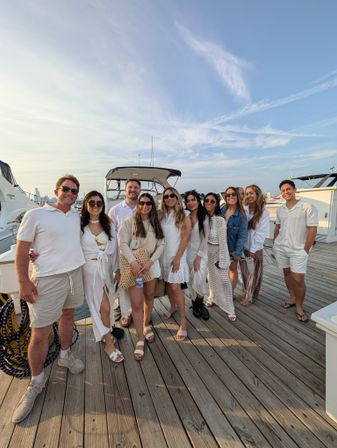 Group of friends in white and neutral summer outfits posing on a wooden marina dock in front of a docked boat, waterfront skyline and wispy blue sky at golden hour.