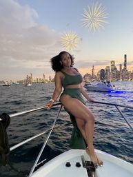 Woman in a green dress posing on a boat bow at sunset with the Chicago skyline, glowing city lights, other boats on the water, and fireworks over Lake Michigan
