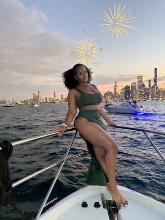 Woman in a green dress posing on a boat bow at sunset with the Chicago skyline, glowing city lights, other boats on the water, and fireworks over Lake Michigan