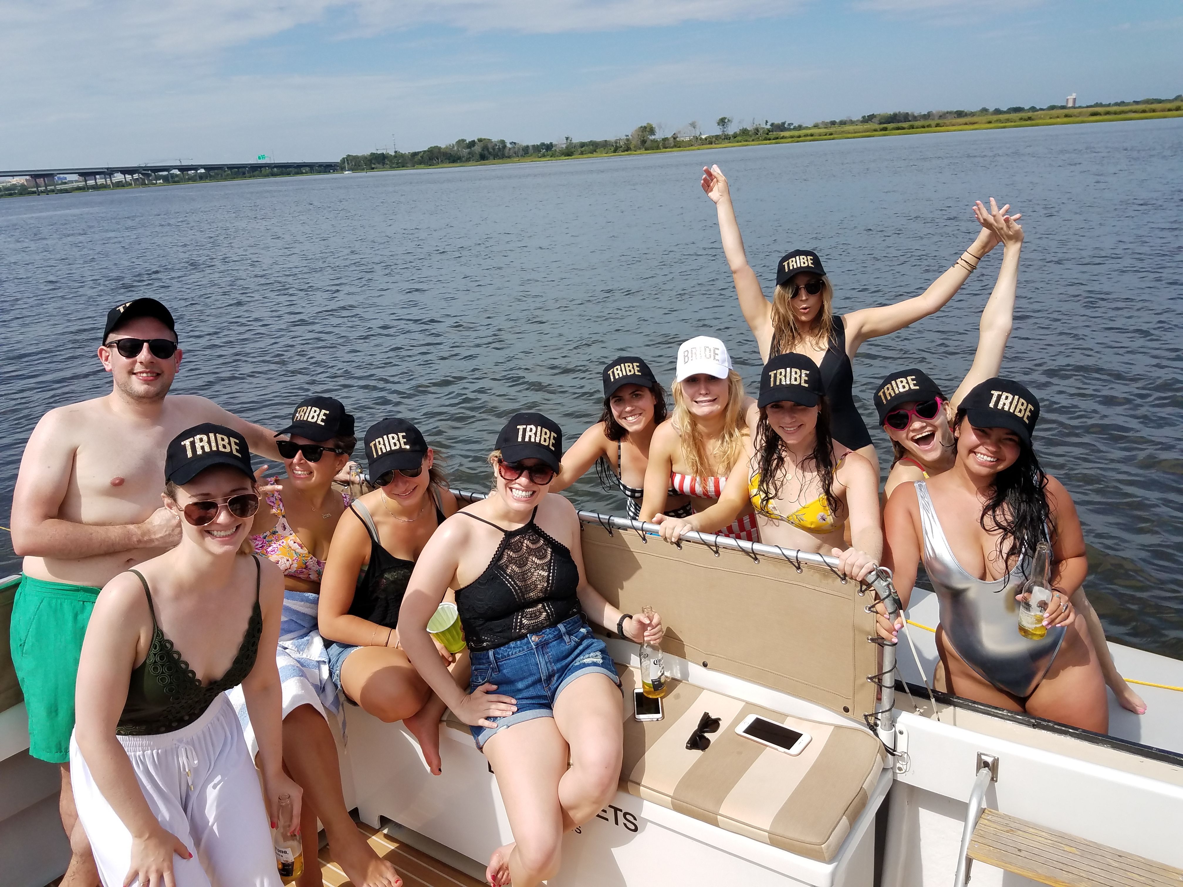 Smiling group of friends in swimsuits wearing matching "TRIBE" caps (one "BRIDE" cap) aboard a sunlit pontoon boat on a calm river with a distant bridge and grassy shoreline.