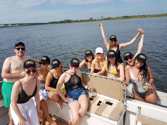 Smiling group of friends in swimsuits wearing matching "TRIBE" caps (one "BRIDE" cap) aboard a sunlit pontoon boat on a calm river with a distant bridge and grassy shoreline.