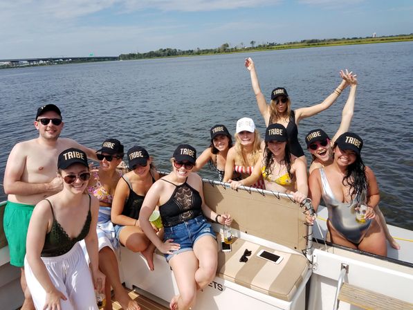 Smiling group of friends in swimsuits wearing matching "TRIBE" caps (one "BRIDE" cap) aboard a sunlit pontoon boat on a calm river with a distant bridge and grassy shoreline.