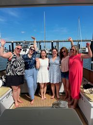 Six smiling women celebrating on a yacht deck at a sunny marina, with sailboats, docks and a bridge under a bright blue sky.