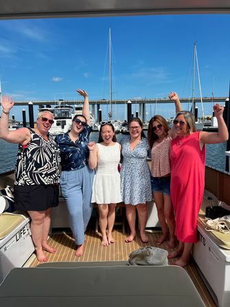 Six smiling women celebrating on a yacht deck at a sunny marina, with sailboats, docks and a bridge under a bright blue sky.