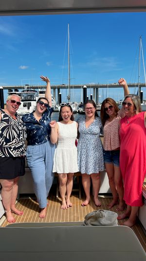 Six smiling women celebrating on a yacht deck at a sunny marina, with sailboats, docks and a bridge under a bright blue sky.