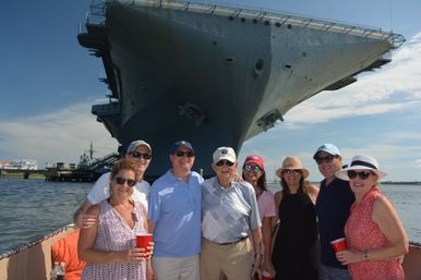 Smiling group of adults on a small boat in a harbor, holding red cups and wearing sunglasses and hats, posing in front of a massive docked aircraft carrier under sunny blue skies.