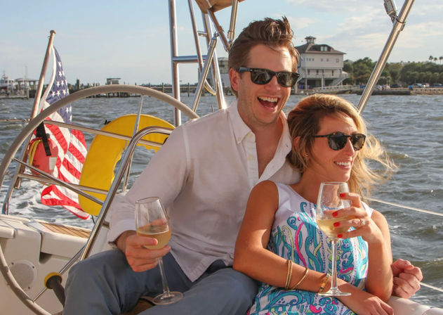 Happy couple in sunglasses sipping white wine on a sailboat near a coastal marina, with the helm, American flag, and waterfront houses in the sunny background.