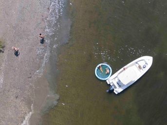 Drone aerial view of a white motorboat anchored in shallow green water off a pebble shoreline, two people lounging on a round inflatable raft tied to the boat and two others walking on the beach.