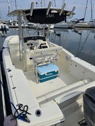 White center-console boat docked at a marina with a stainless T-top and rod holders, helm and steering wheel visible, blue cooler secured under the leaning post, calm harbor water and sailboats in the background.