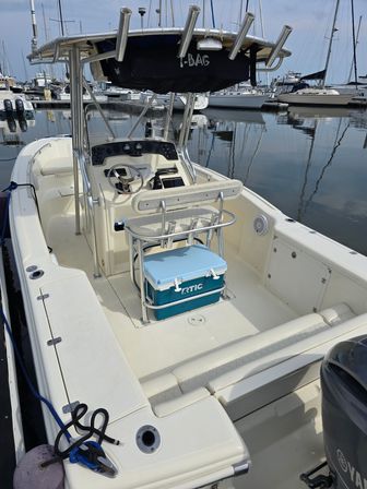 White center-console boat docked at a marina with a stainless T-top and rod holders, helm and steering wheel visible, blue cooler secured under the leaning post, calm harbor water and sailboats in the background.