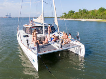 Group of people sunbathing and socializing on the trampoline and deck of a white sailing catamaran anchored in a calm coastal bay near a sandy beach and tree-lined shoreline on a sunny day.