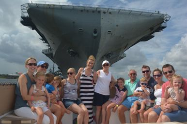 Smiling multigenerational family and children seated on a boat, posing in front of the enormous bow of an aircraft carrier docked at a harbor under a dramatic cloudy sky.