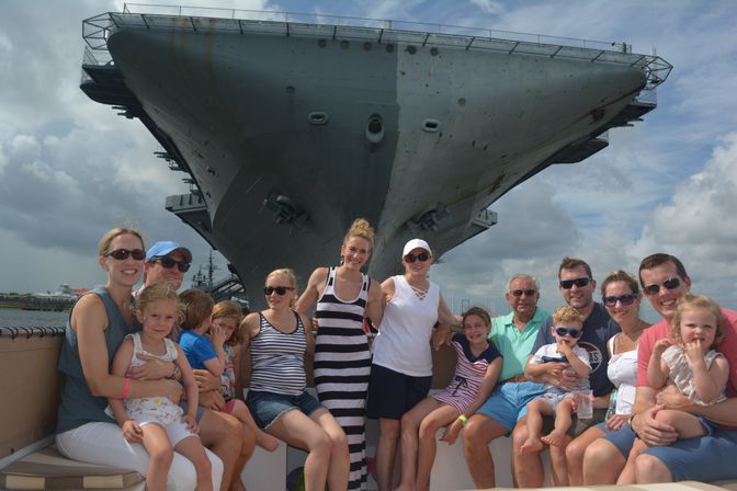 Smiling multigenerational family and children seated on a boat, posing in front of the enormous bow of an aircraft carrier docked at a harbor under a dramatic cloudy sky.