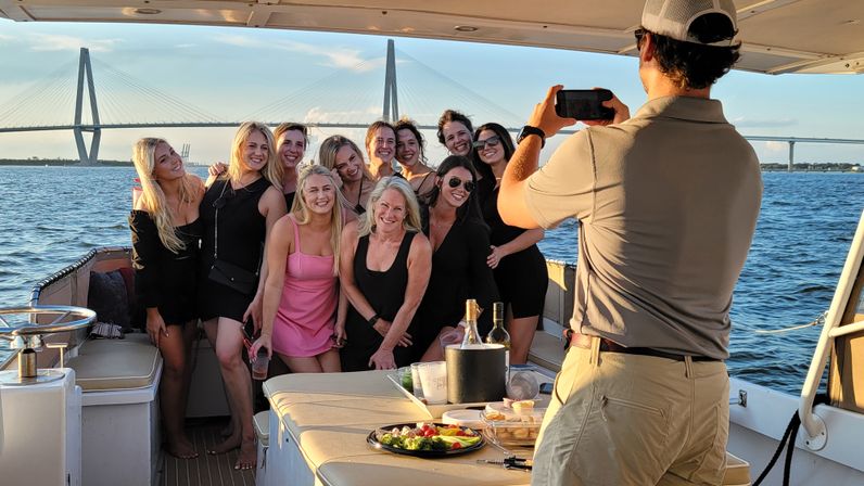 Group of women posing for a sunset yacht cruise photo, drinks and appetizer platter on the deck, blue water and a cable-stayed bridge spanning the coastal skyline in the background.