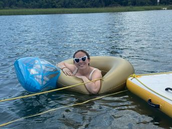 Smiling person wearing heart-shaped sunglasses relaxing in a gold inflatable tube on a calm lake, tethered to a blue float and yellow paddleboard with grassy, tree-lined shore in the background.