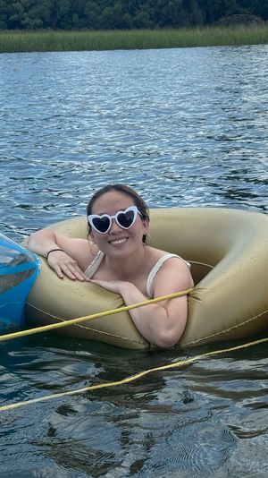 Smiling person wearing heart-shaped sunglasses relaxing in a gold inflatable tube on a calm lake, tethered to a blue float and yellow paddleboard with grassy, tree-lined shore in the background.