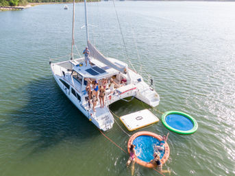Aerial view of a white catamaran anchored in a sunny coastal bay with friends on deck and colorful inflatable water trampolines and floating pads nearby